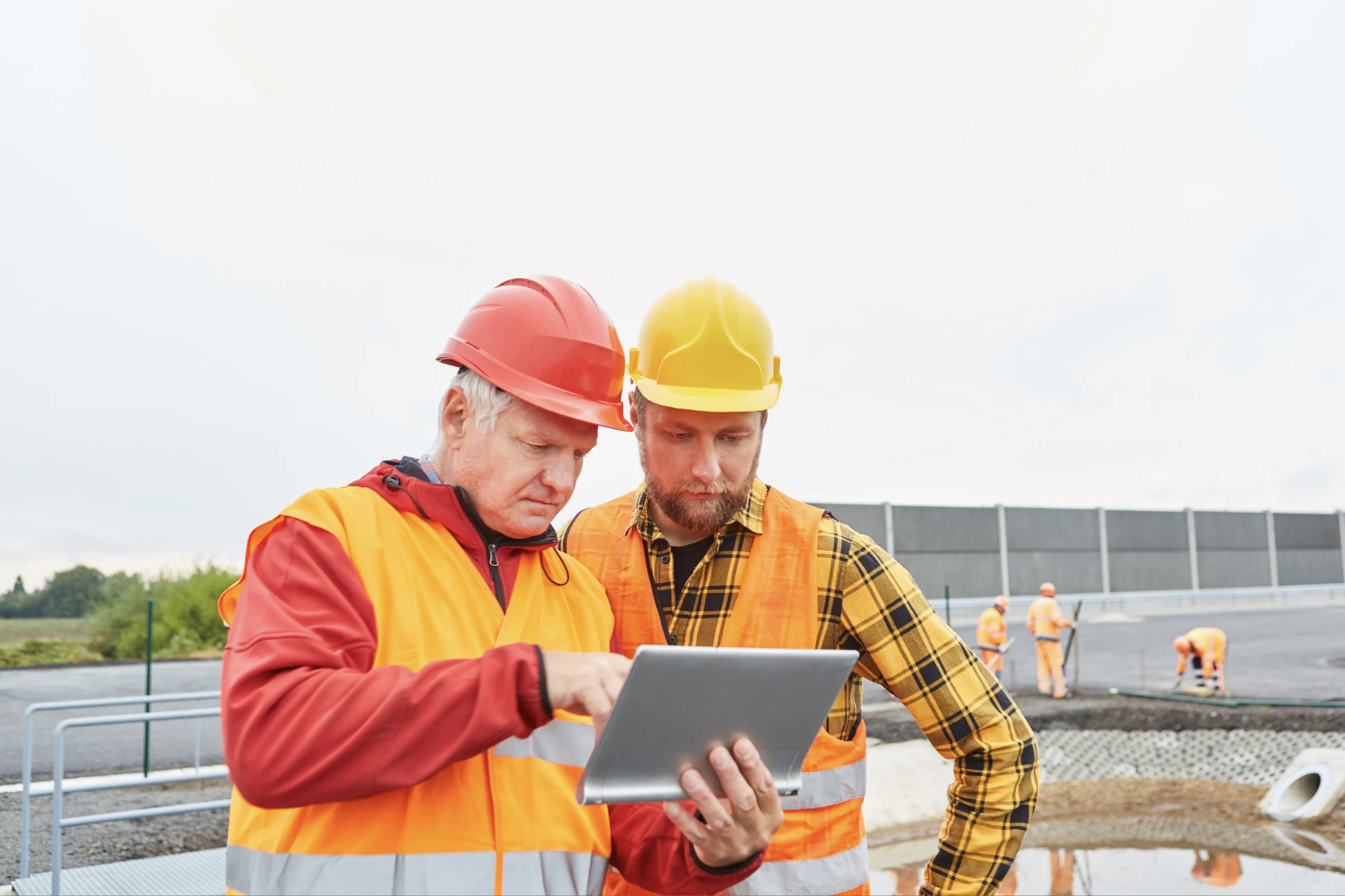 Two construction workers in hard hats and safety vests looking at a tablet together